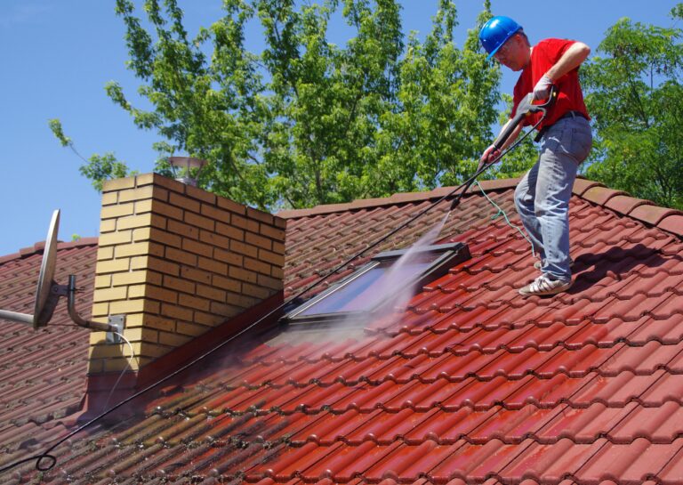 A worker in a blue hard hat and red shirt uses a pressure washer to clean red roof tiles on a house. The chimney and a satellite dish.