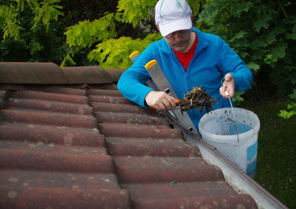 A person wearing a cap and blue jacket is using a trowel to remove debris from a gutter, holding a bucket nearby, surrounded by green foliage.