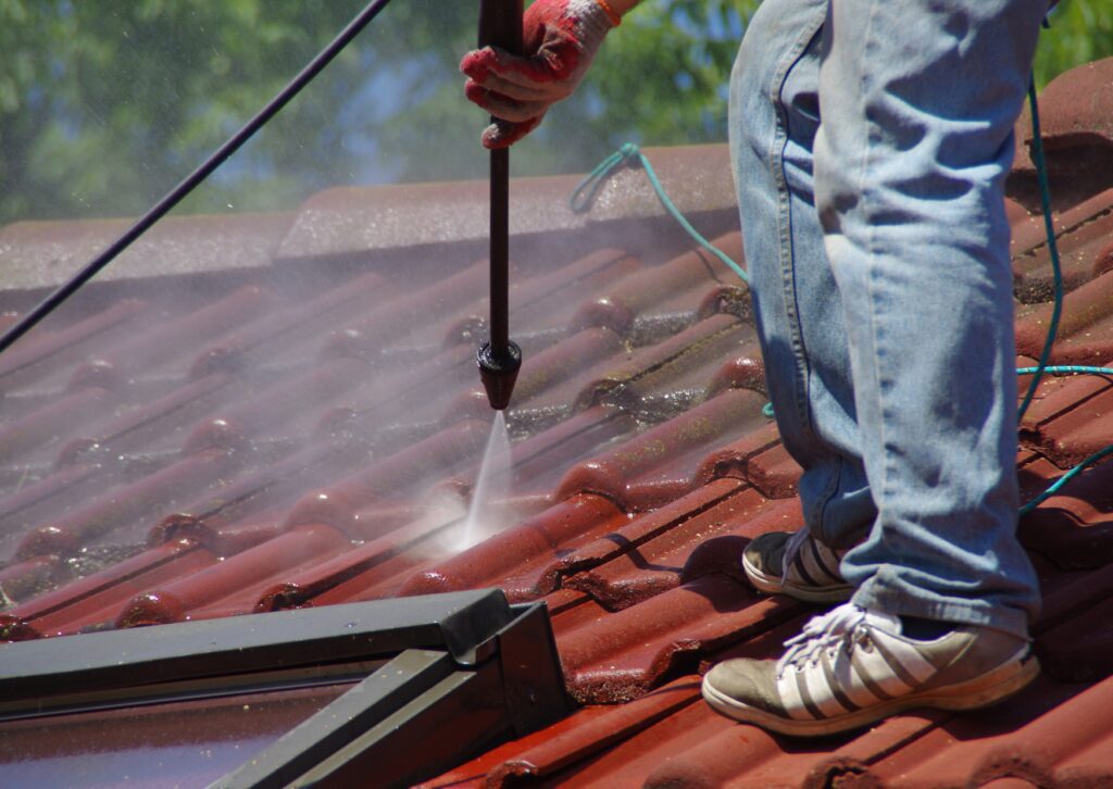A person wearing gloves and sneakers is using a high-pressure washer to clean red terracotta roof, standing on the roof while water sprays.