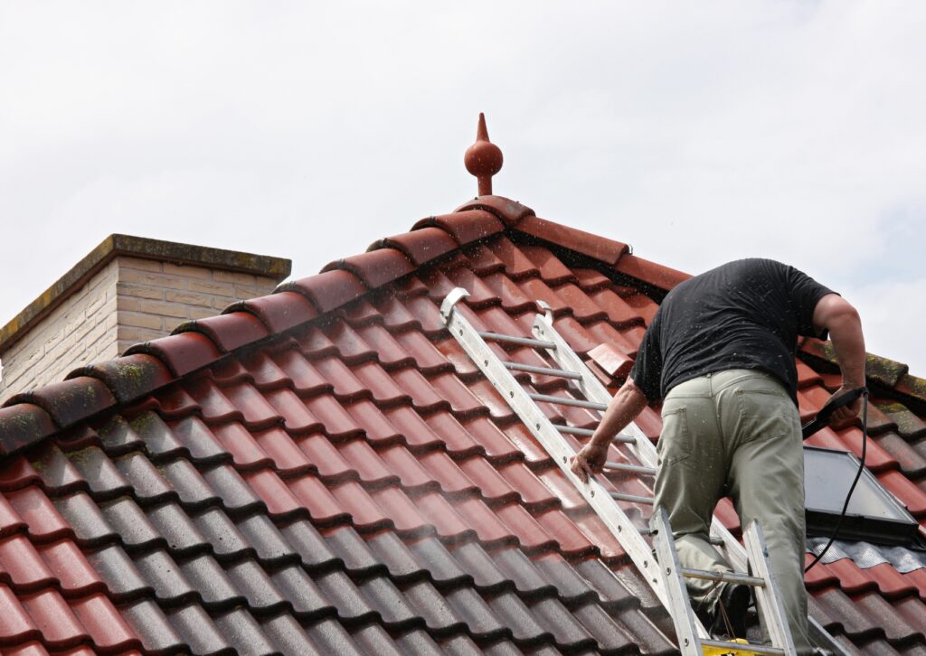 A worker stands on a ladder, pressure washing red and gray canopy tiles on a house roof. The roof features a decorative finial at the peak.