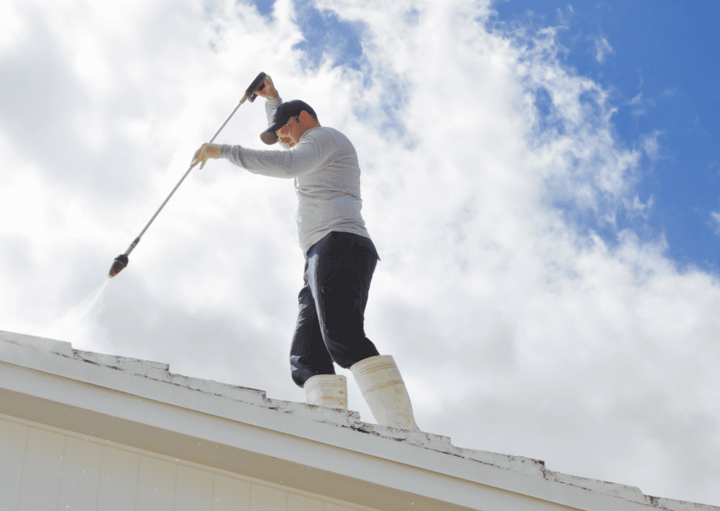 A worker cleaning a roof using a power washer, demonstrating the process of soft washing your roof under a bright sky.