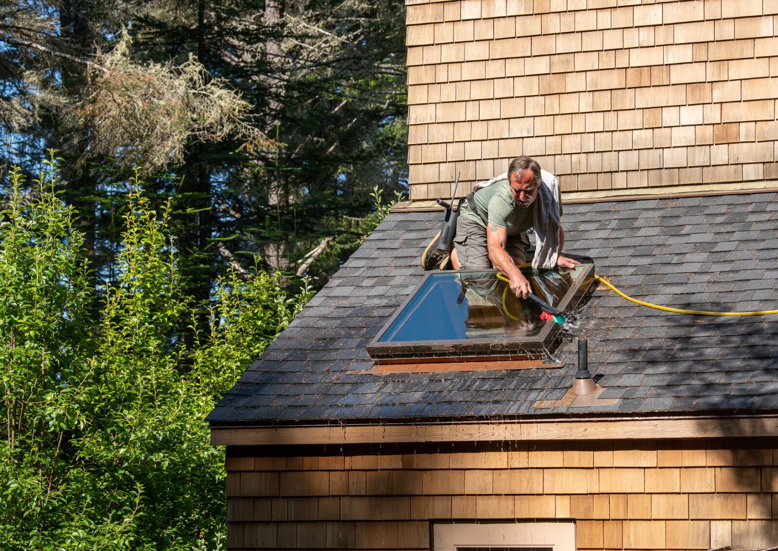Man performing soft wash roof cleaning around a skylight using a hose and brush on asphalt shingles.