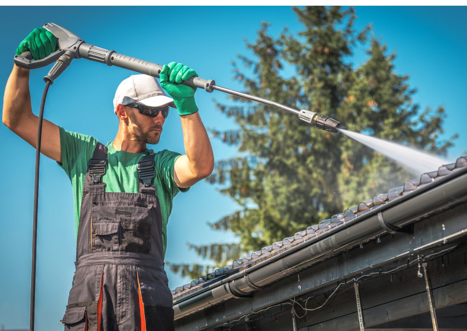 A man using a sprayer for soft wash roof cleaning on shingles, wearing gloves and safety gear outdoors.