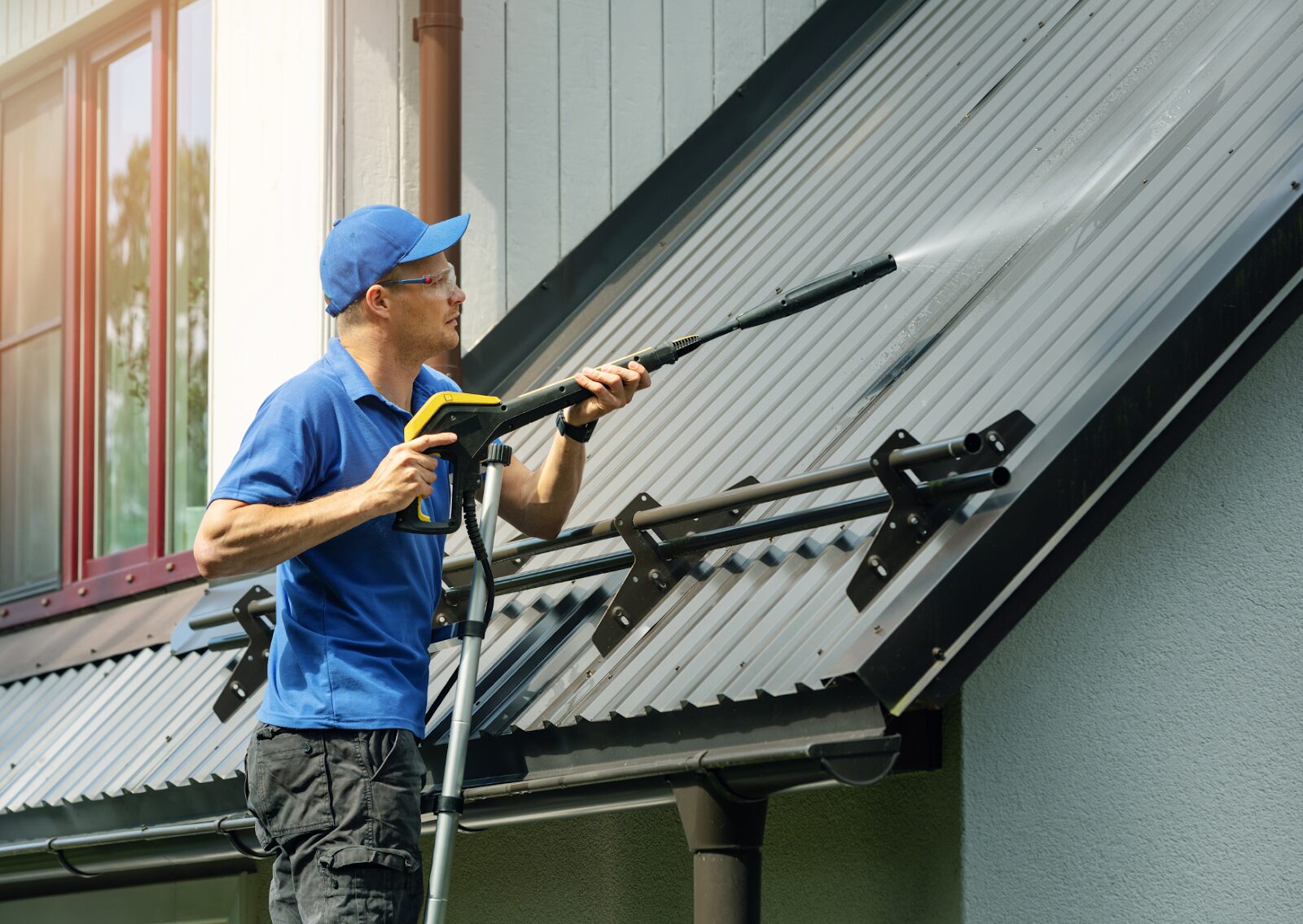 A worker spraying a metal roof with water, demonstrating what is softwashing a roof to remove buildup safely.