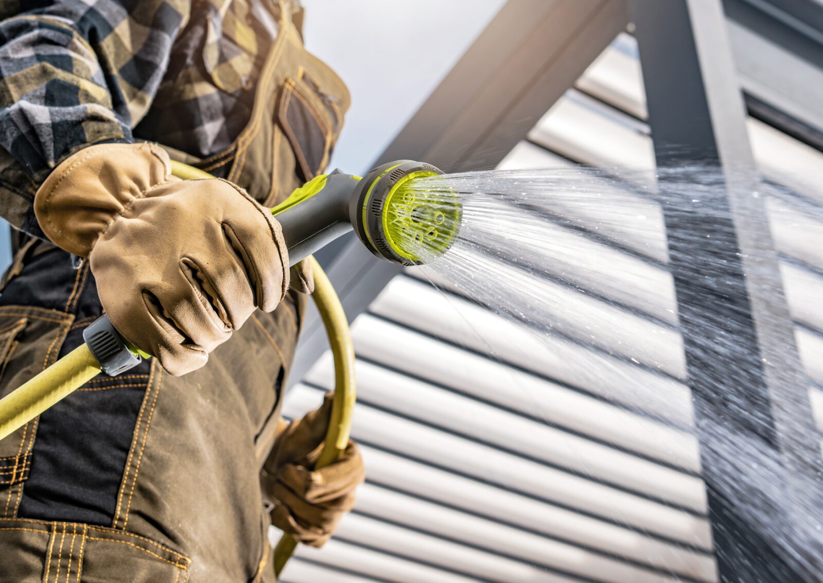 Worker in gloves and overalls spraying water with garden hose nozzle for outdoor cleaning or rinsing.
