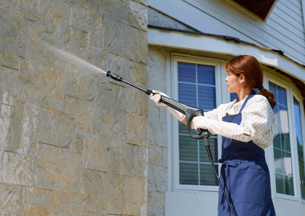 Woman spraying exterior wall with pressure washer to show the benefits of soft washing your house effectively.