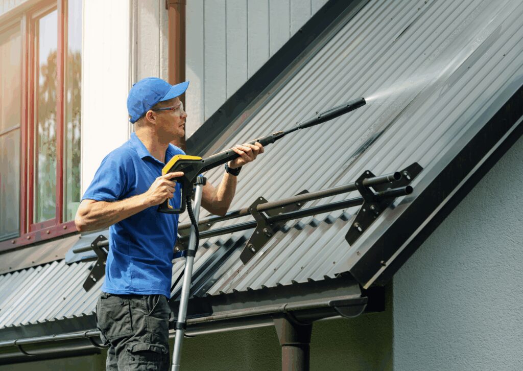 Technician on ladder performs roof soft washing on a metal roof, using a pressure wand to rinse away dirt and grime.