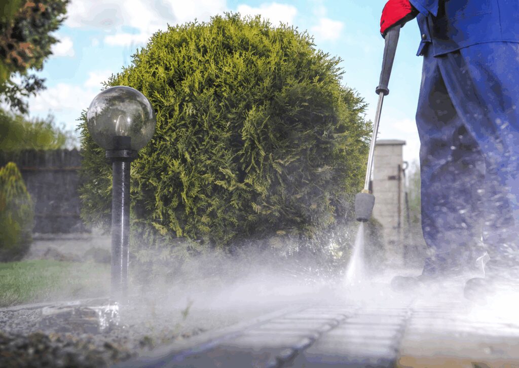 Worker pressure washing a garden pathway with water spray near landscaping and an outdoor lamp post.
