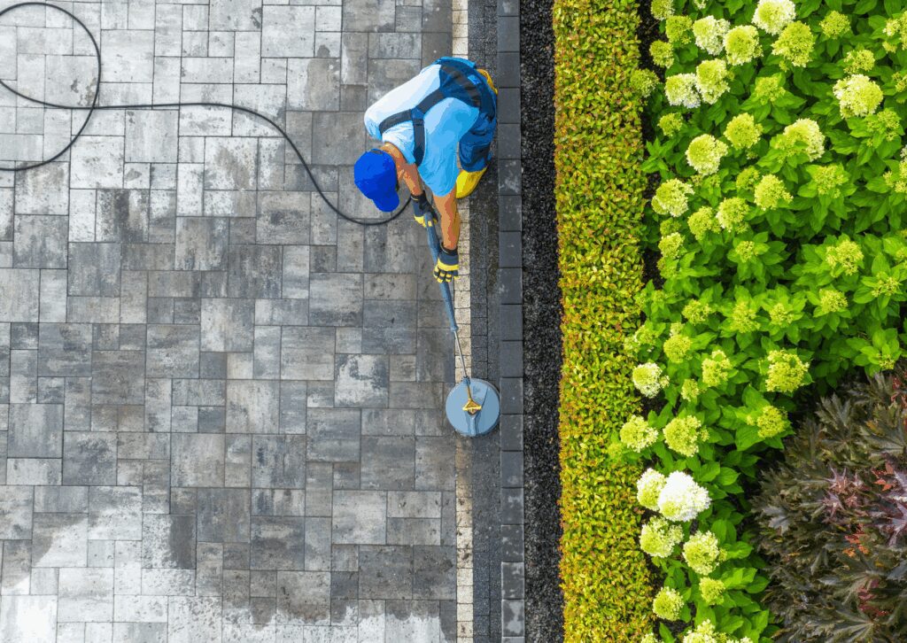 Top view of a worker using a surface cleaner to pressure wash a stone patio next to a colorful garden bed.