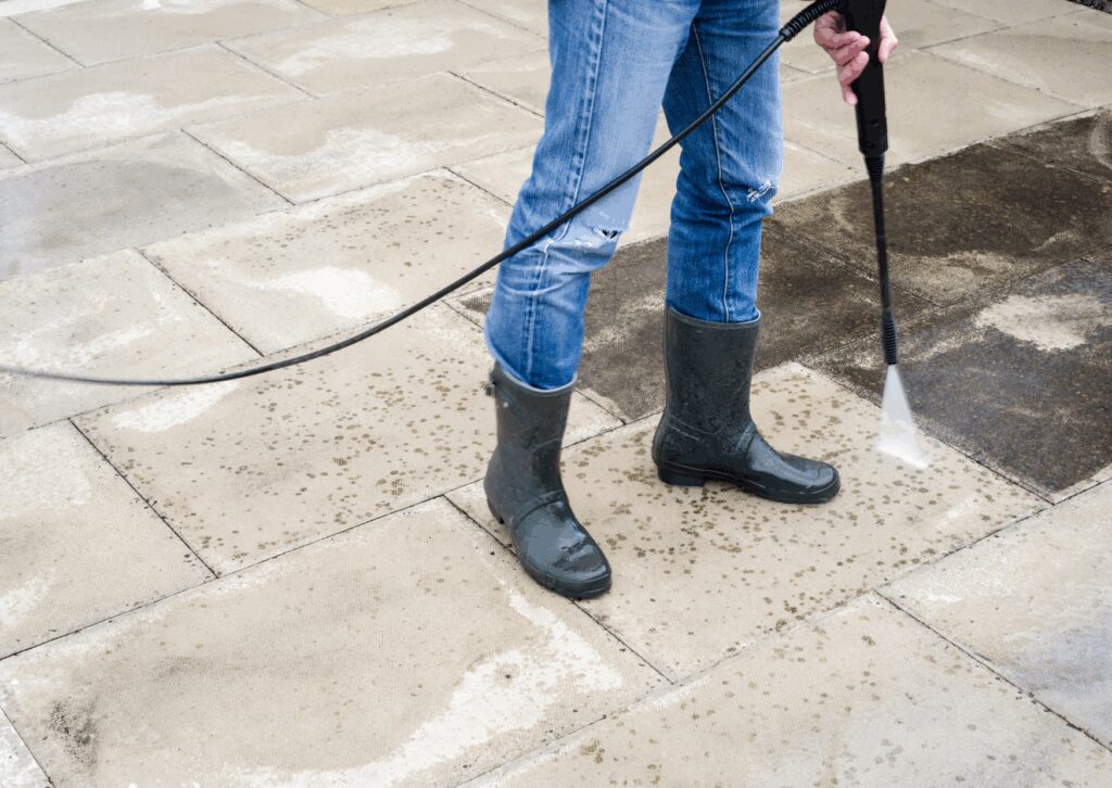 Person in jeans and rubber boots using a soft washing system to clean dirty patio paving slabs with high pressure spray.